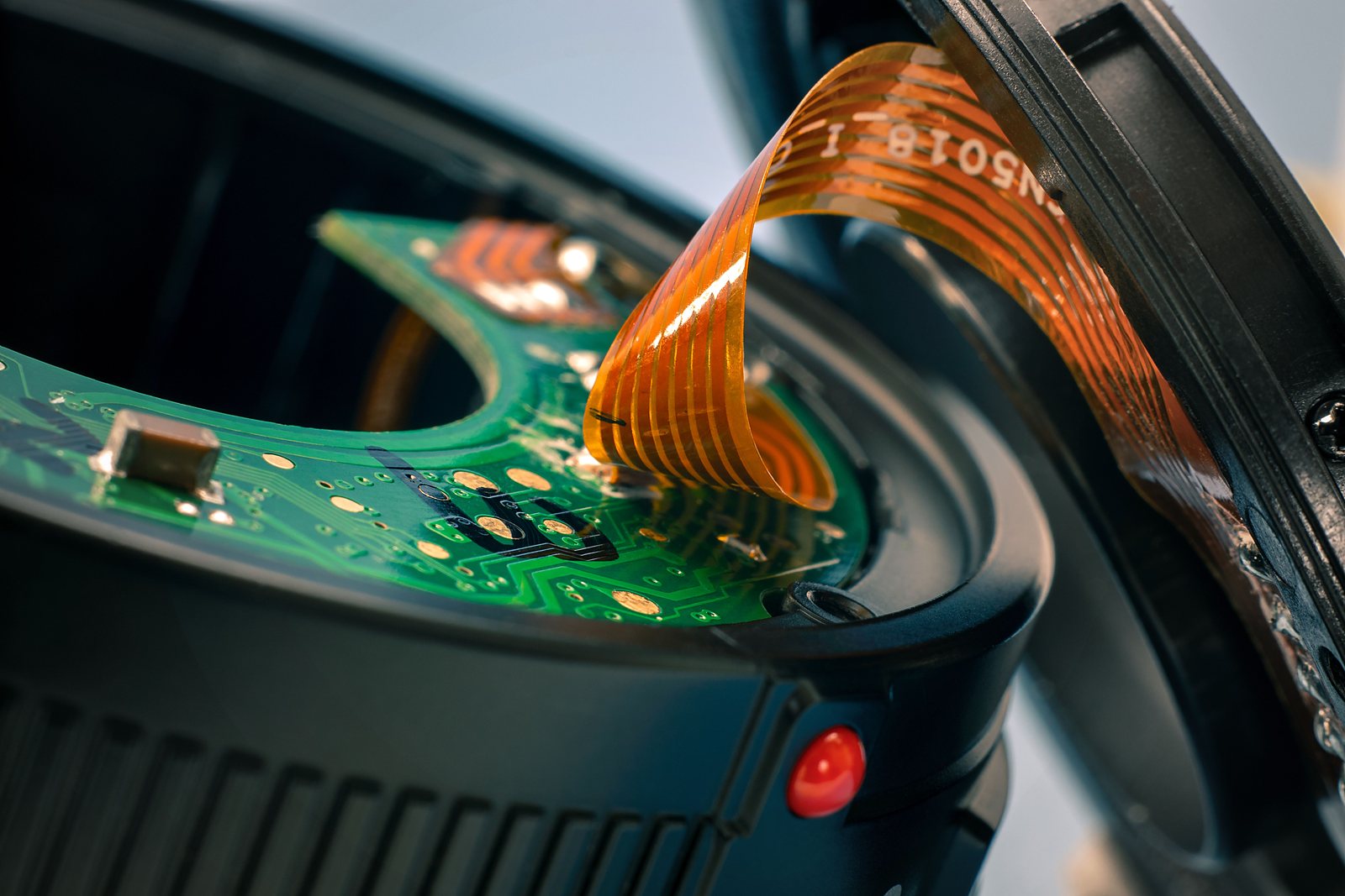 Close-up of a populated green board: a small passive beside exposed gold pads and a polyimide flex tail